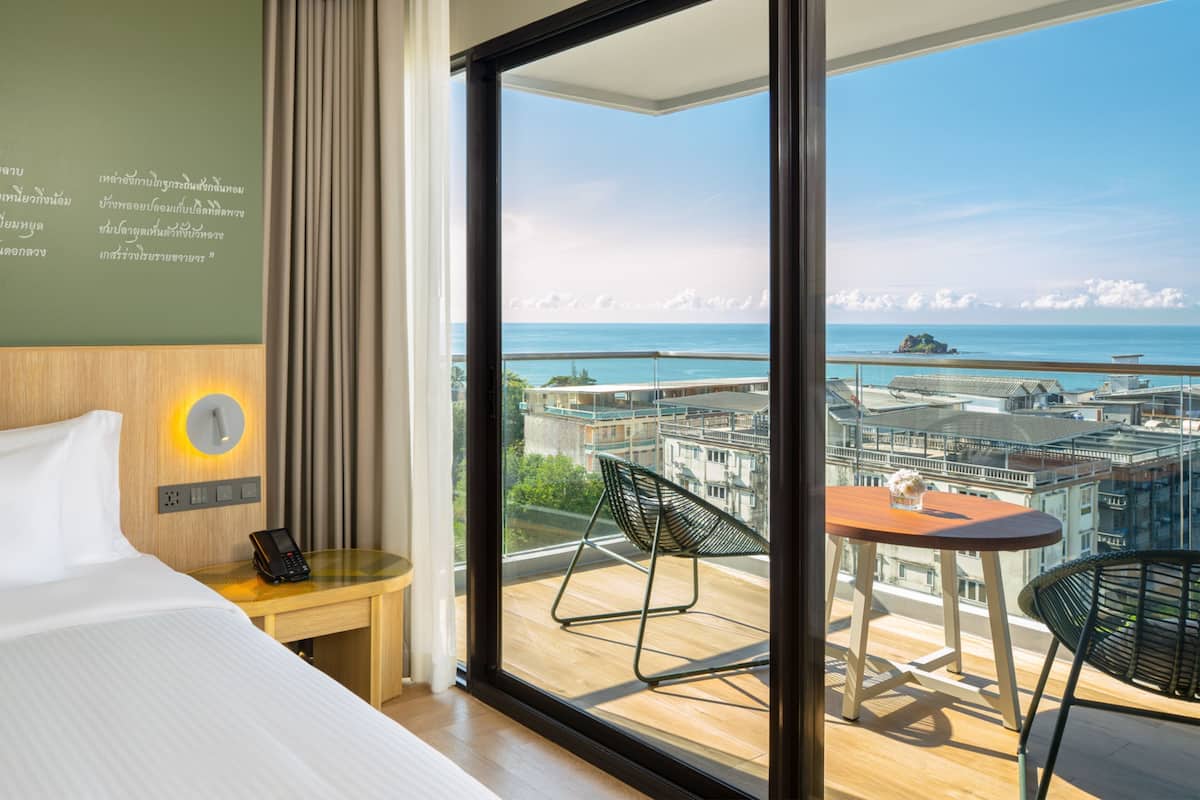 A hotel room with a large window, a bed, a bedside table with a phone, and a view of a cityscape and ocean.