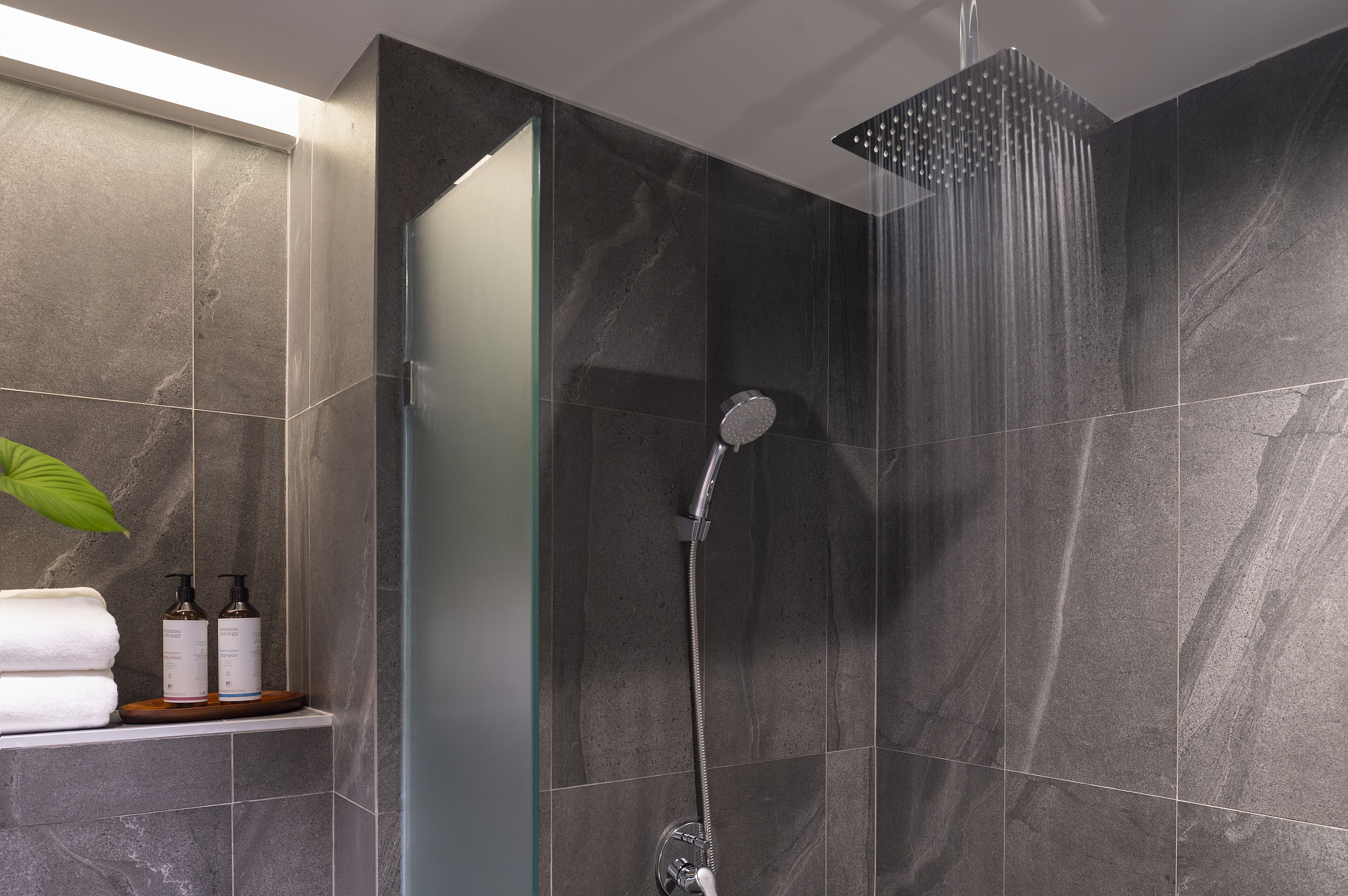 A modern bathroom with dark marble tiles, a glass shower enclosure, and a shelf with toiletries.