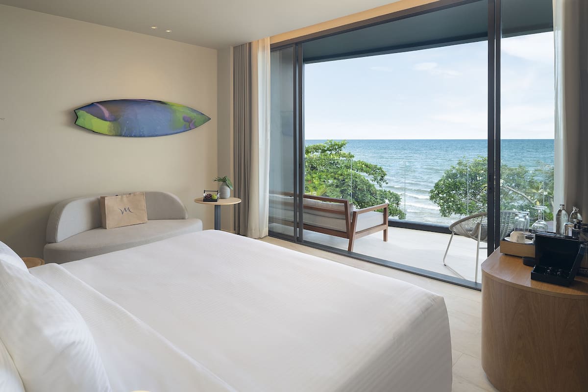 A modern hotel room with a large bed, a seating area with a view of the sea, and a small table with a plant and a bag.