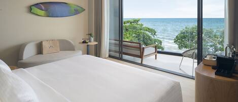 A modern hotel room with a large bed, a seating area with a view of the sea, and a small table with a plant and a bag.
