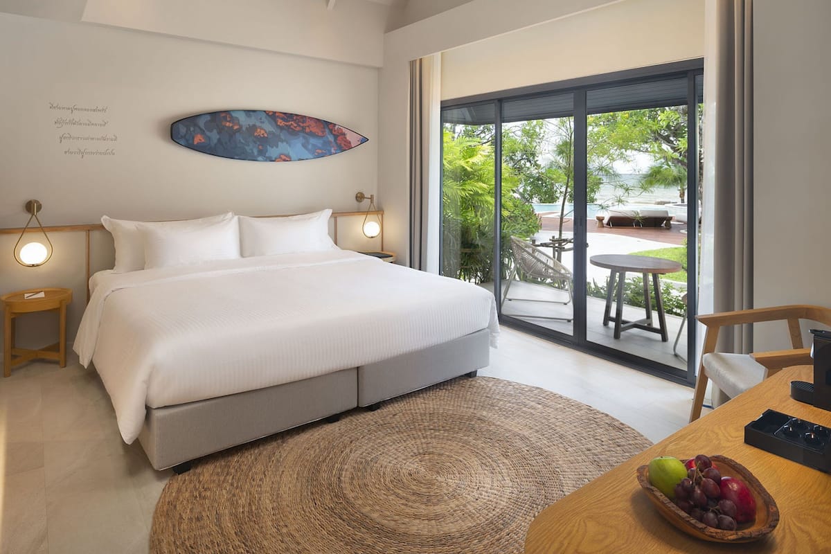 A modern bedroom with a large bed, a view of the outdoors, and a dining area with a fruit bowl.