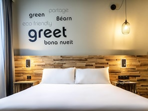 A hotel room with a wooden headboard, a bed with white bedding, and a wall with a light fixture and a word cloud.