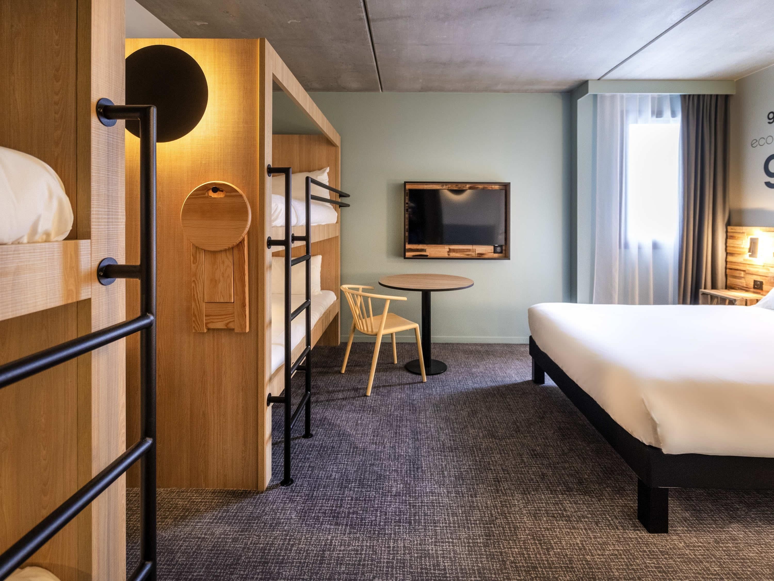 Una habitación de hotel con una cama litera, una cama individual, un televisor, una mesita pequeña y una silla.