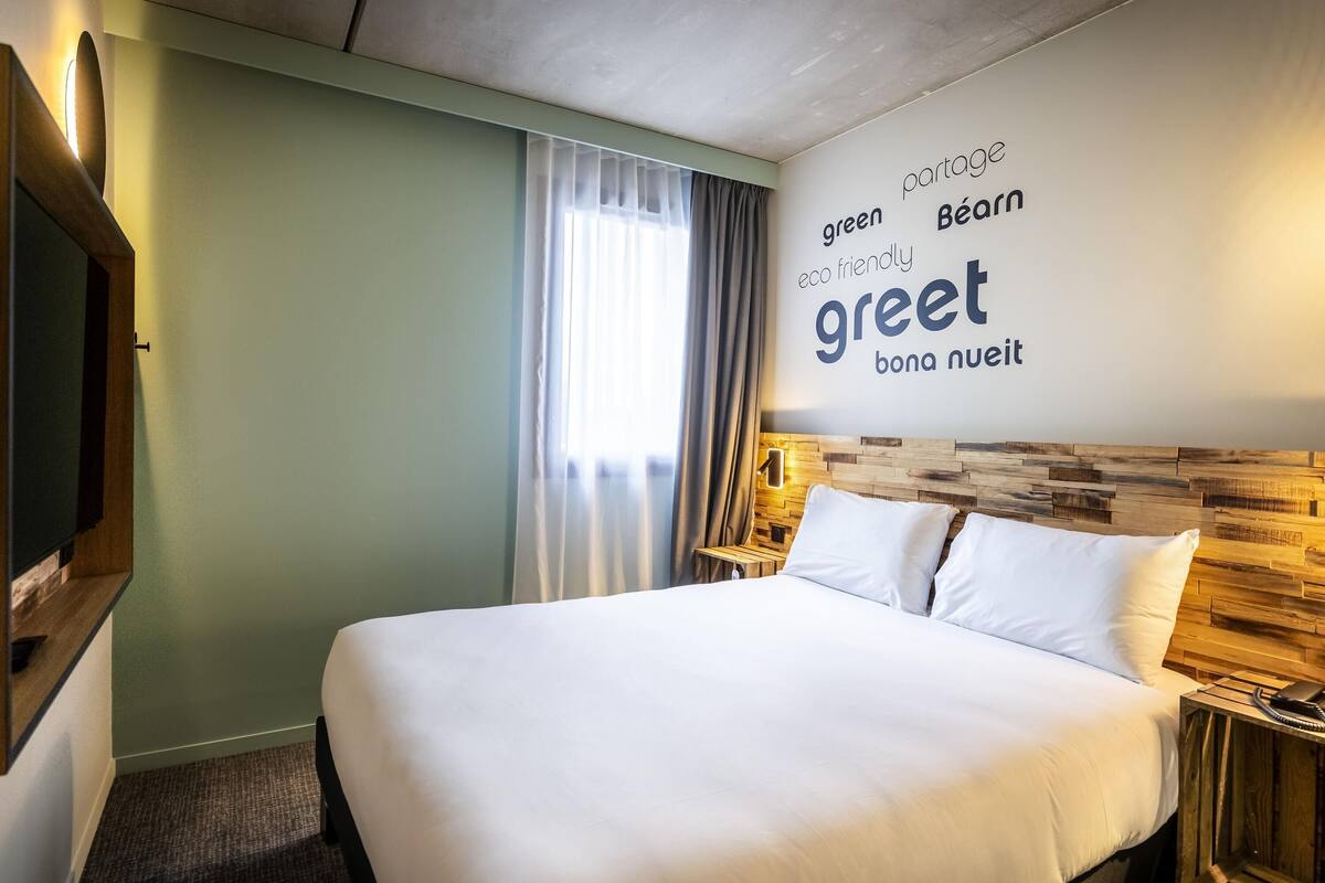 Une chambre d’hôtel avec un lit, une table de chevet, une horloge murale, un miroir et une fenêtre avec des rideaux.