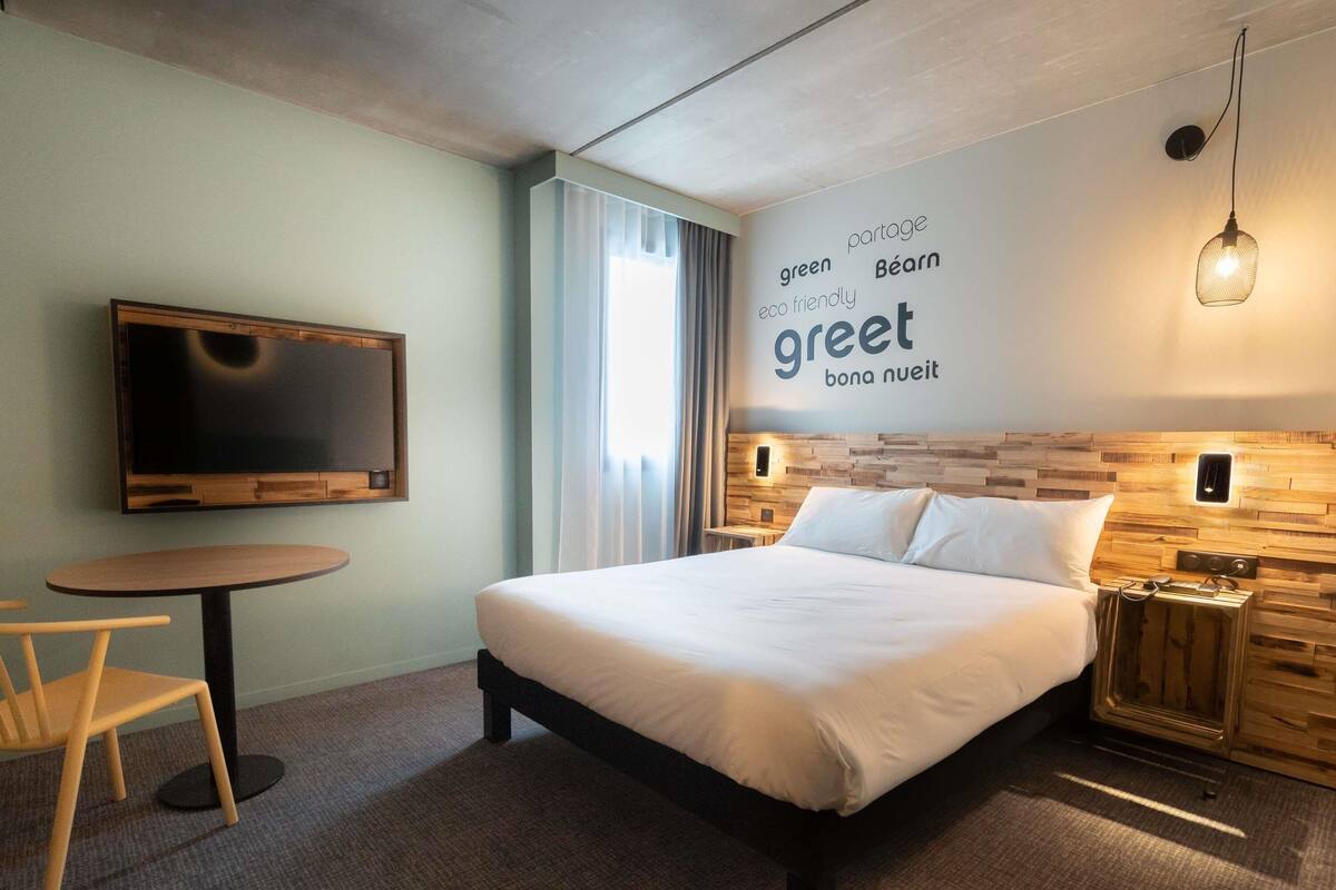 Une chambre d’hôtel moderne dotée d’un grand lit, d’un téléviseur à écran plat, d’une table ronde et d’un mur décoré dans un style écologique et respectueux de l’environnement.