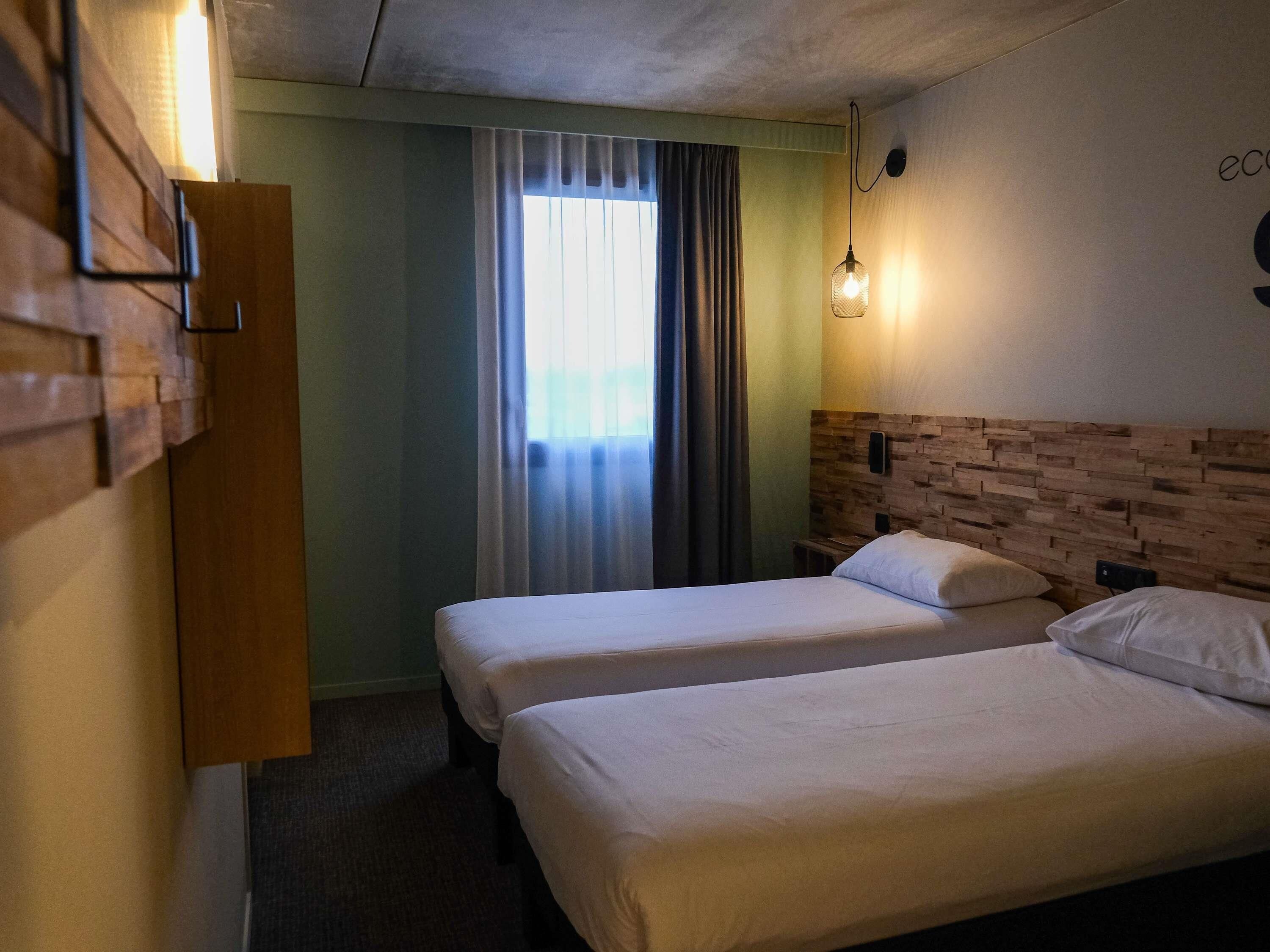 Et hotelværelse med to senge, en træsenggavl, en vægmonteret lampe og et vindue med gardiner.