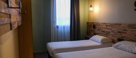 Habitación de hotel con dos camas individuales, cabecero de madera, luz de pared y ventana con cortinas.