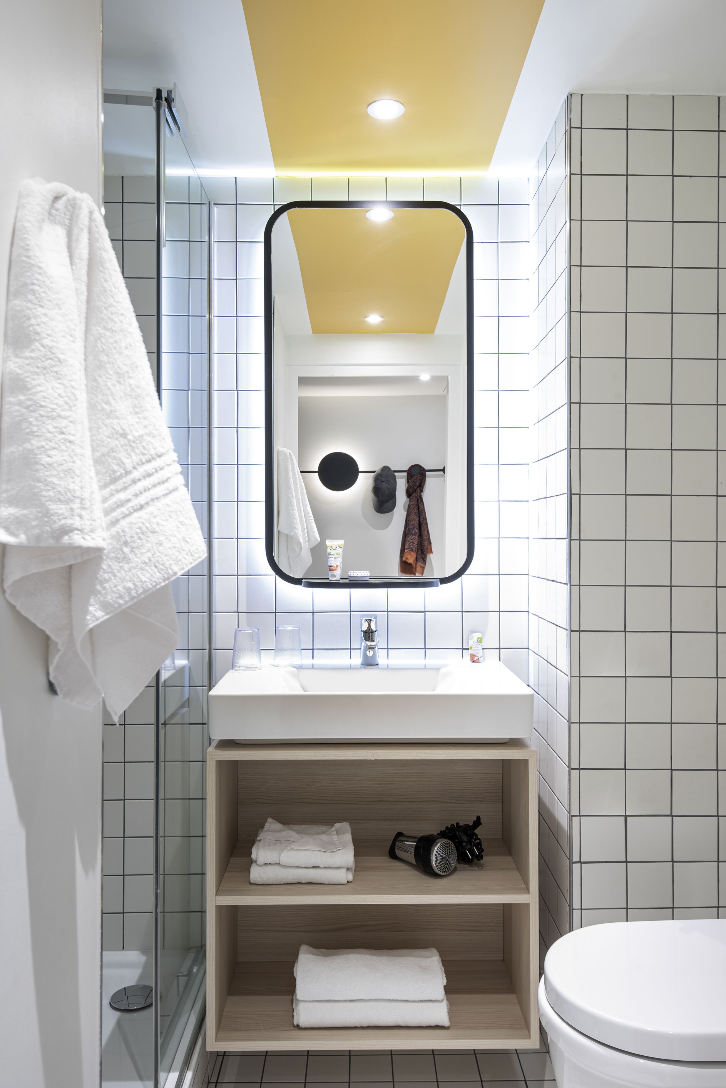 Un baño moderno con un lavamanos, espejo y retrete blancos. Hay repisas con toallas y un secador de pelo.