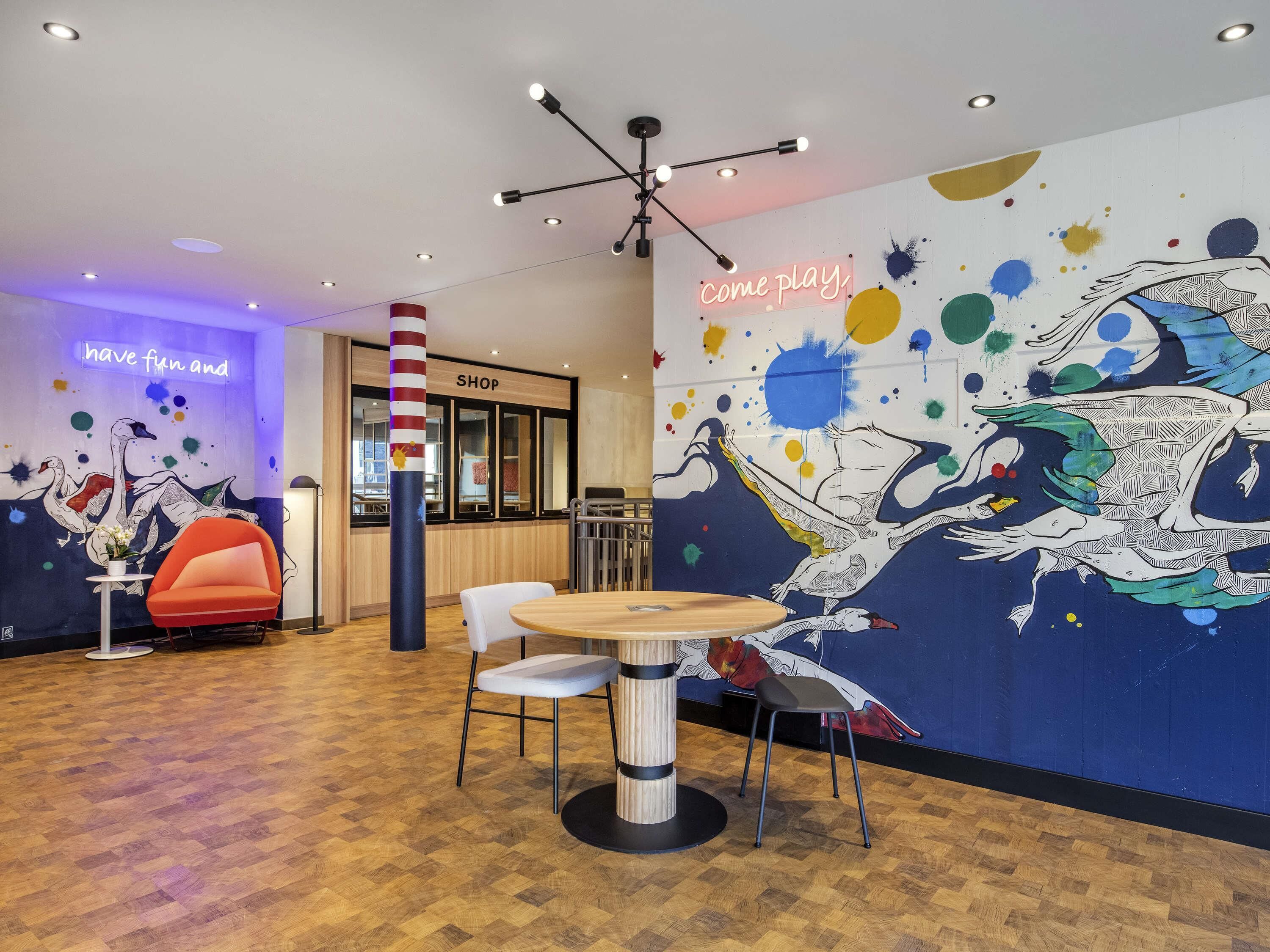 A modern interior with a wooden floor, a round table with chairs, a colorful mural, and a neon sign.