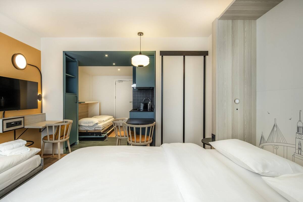 A hotel room with a large bed, a desk, a chair, a small kitchenette, and a television.