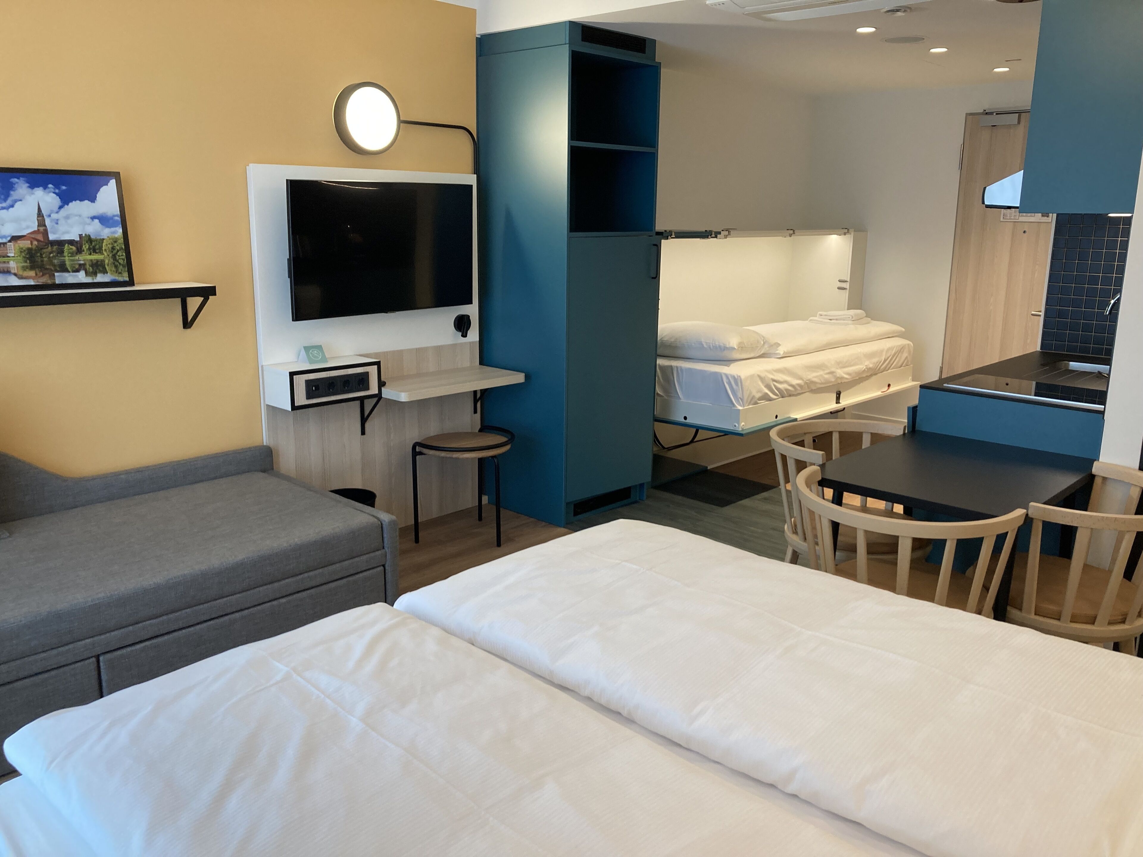A modern hotel room with a bed, a desk, a chair, a TV, and a small stool.