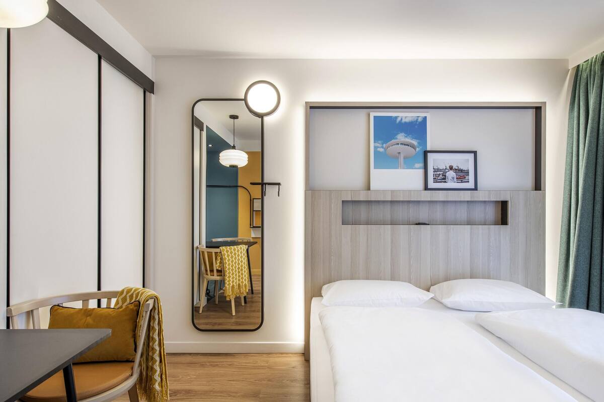 A hotel room with a bed, a desk, a chair, a mirror, and a wall-mounted shelf with pictures.