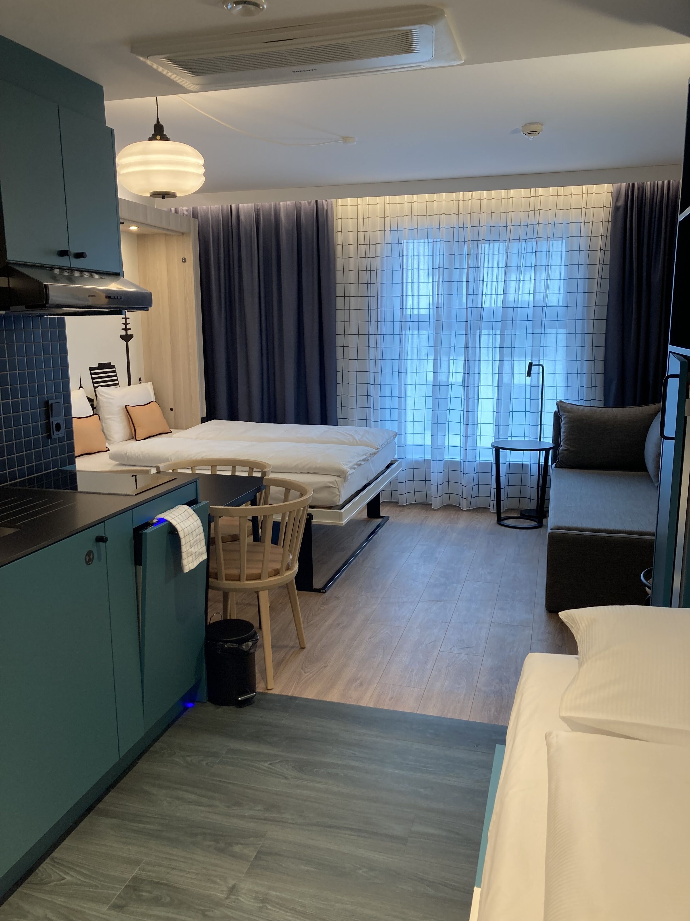 A compact hotel room with a kitchenette, a bed, a seating area, and a window with curtains.