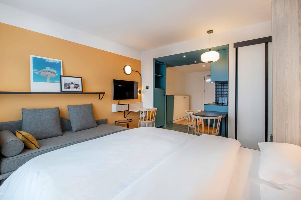 A modern hotel room with a large bed, a sofa, a desk with a chair, a television, and a kitchen area visible through an open door.