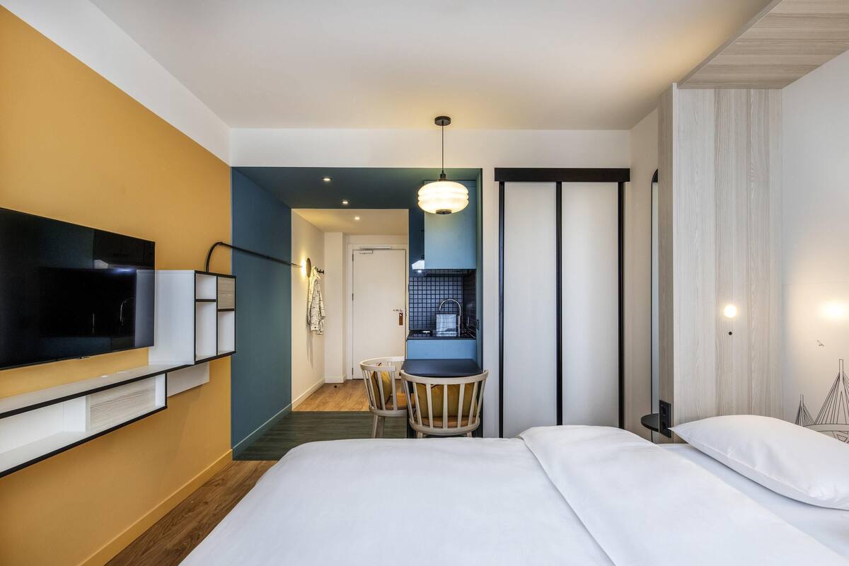 A hotel room with a bed, a TV, a desk with a chair, and a kitchenette.