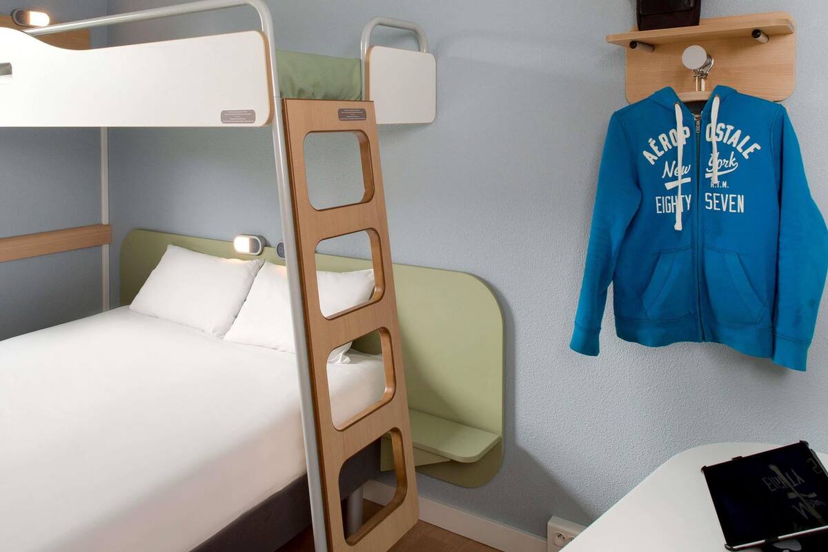 Un lit superposé avec une échelle en bois, une table de chevet sur laquelle repose un livre, et une veste bleue accrochée à un crochet.