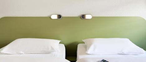 Habitación de hotel con dos camas, cada una con ropa de cama y almohadas blancas. Hay una lámpara de pared sobre las camas y una pared verde con diseño que imita pasto.