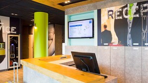 A reception desk with a computer monitor, a green pillar, and a wall with a poster. - ibis budget Montceau-les-Mines (Montceau-les-Mines)