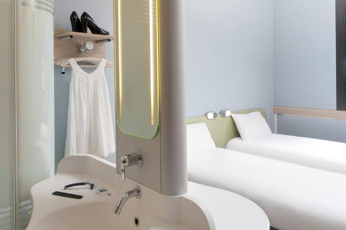 Une chambre d’hôtel avec un lavabo blanc, un miroir, un lit avec une literie blanche et une étagère avec des chaussures.