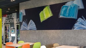 A modern cafe with colorful chairs and tables, a mural of books on the wall, and a vending machine labeled 'COOL SERVICE'. - ibis budget Montceau-les-Mines (Montceau-les-Mines)