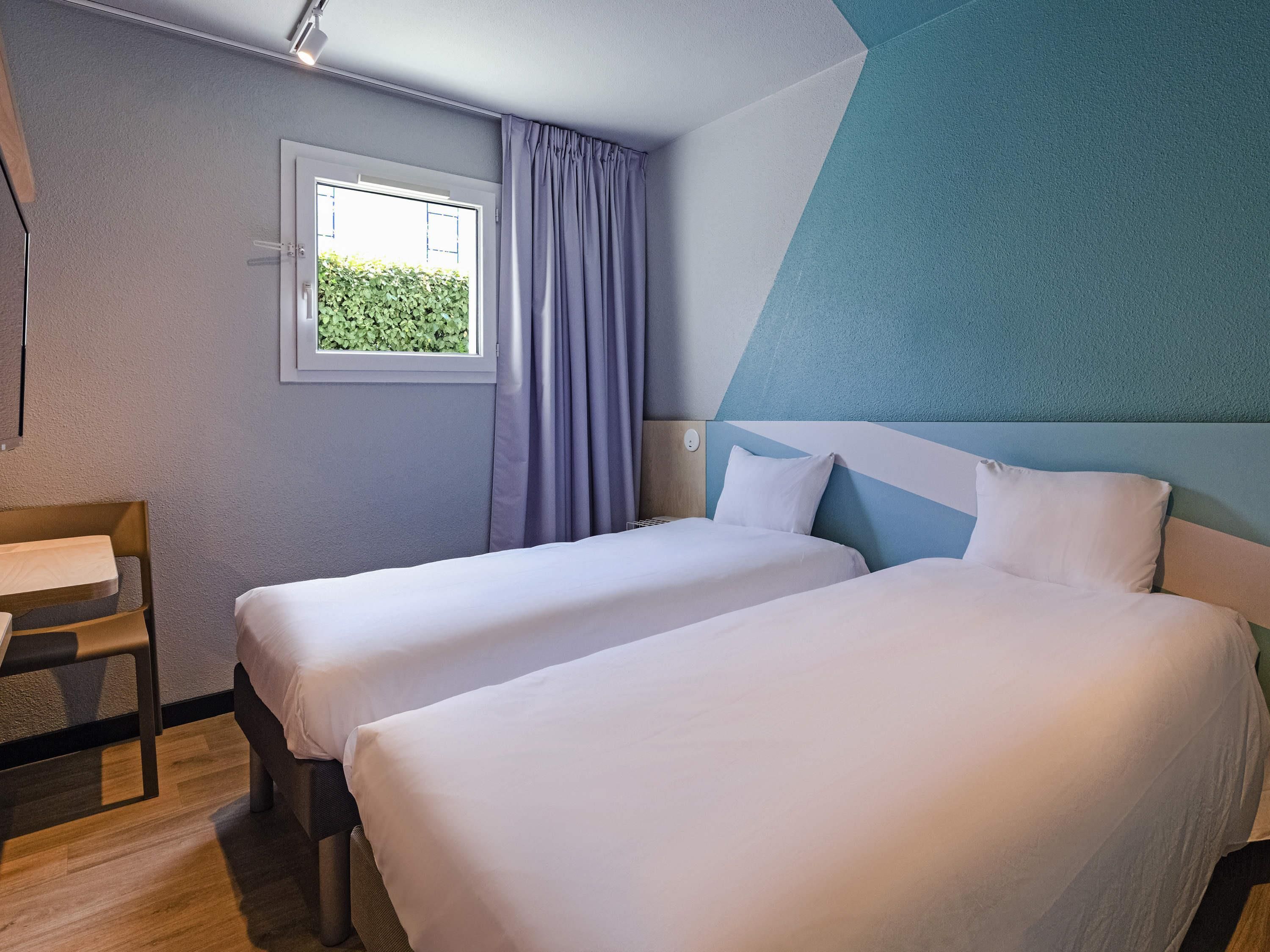 Une chambre d’hôtel avec deux lits, une fenêtre avec des rideaux, un bureau et une vue sur la verdure.