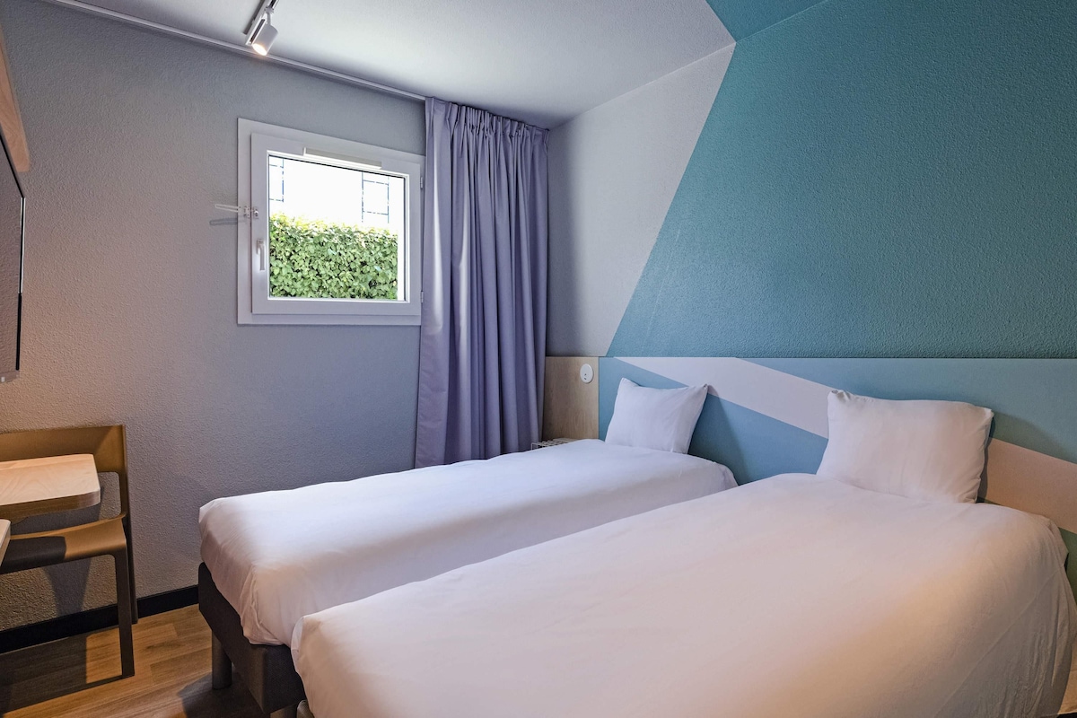 Hotellihuone, jossa on kaksi sänkyä, ikkuna verhoilla, työpöytä ja näkymä vehreään maisemaan.