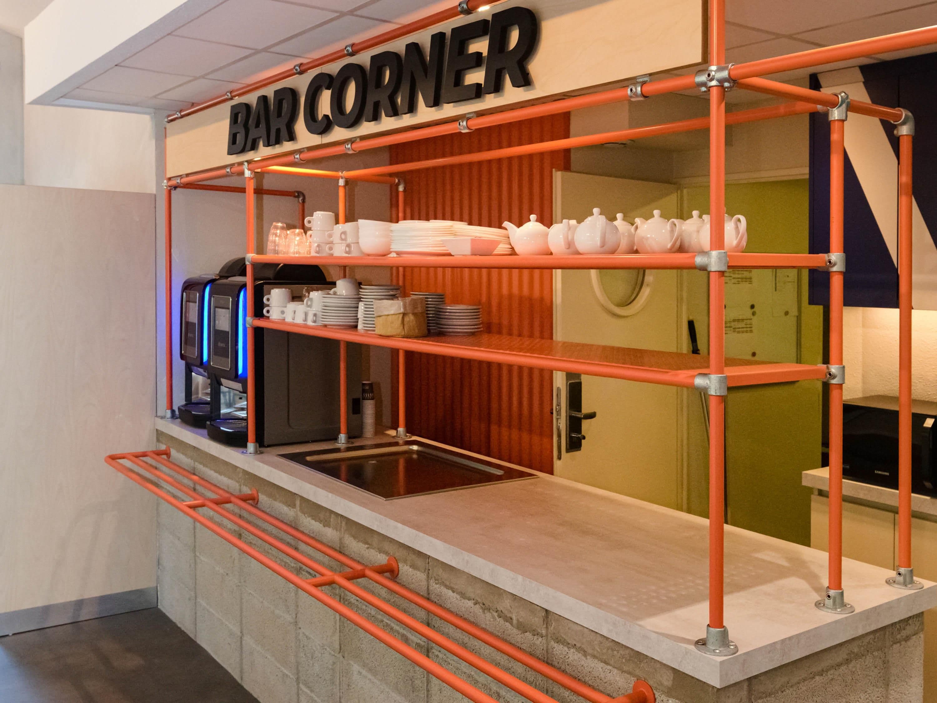 Un espace bar agrémenté d’une enseigne indiquant « BAR CORNER », équipé d’une machine à café, d’étagères remplies d’assiettes et d’un comptoir.