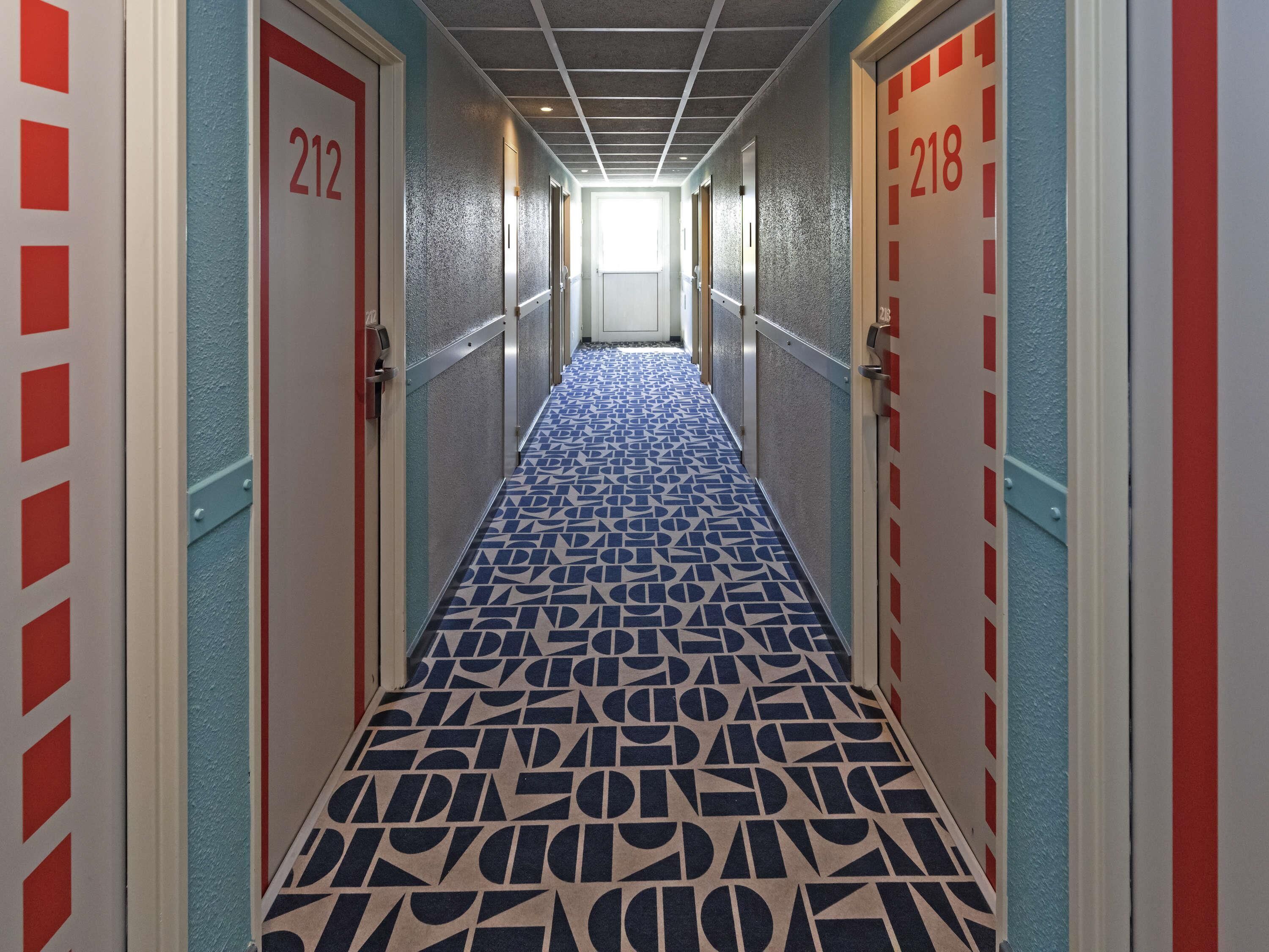 Un couloir avec un tapis à motifs, des portes numérotées 212 et 218, et un plafond orné d’un motif en réseau.