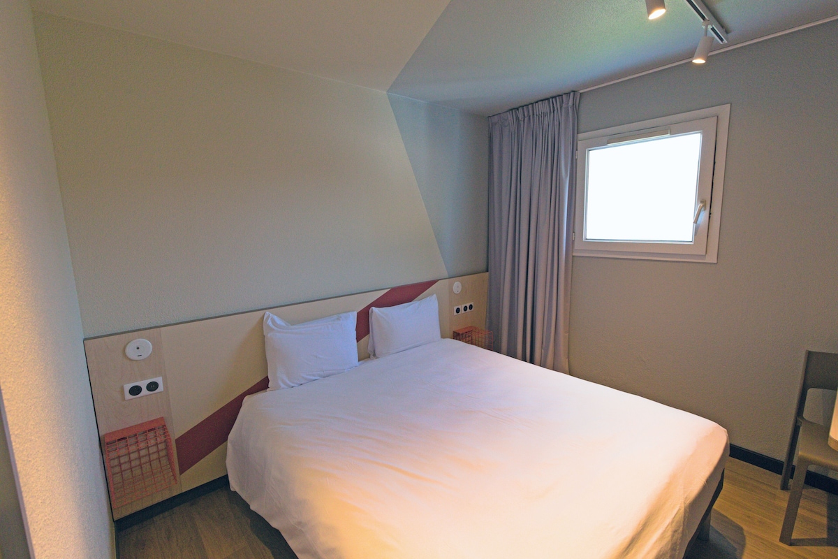 Una habitación de hotel con cama, ventana con cortinas, lámpara de pared y una pequeña cesta roja.