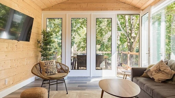 A wooden interior with a TV mounted on the wall, a wicker chair, a sofa, and a glass door leading outside. - River House Getaway (Vienna)