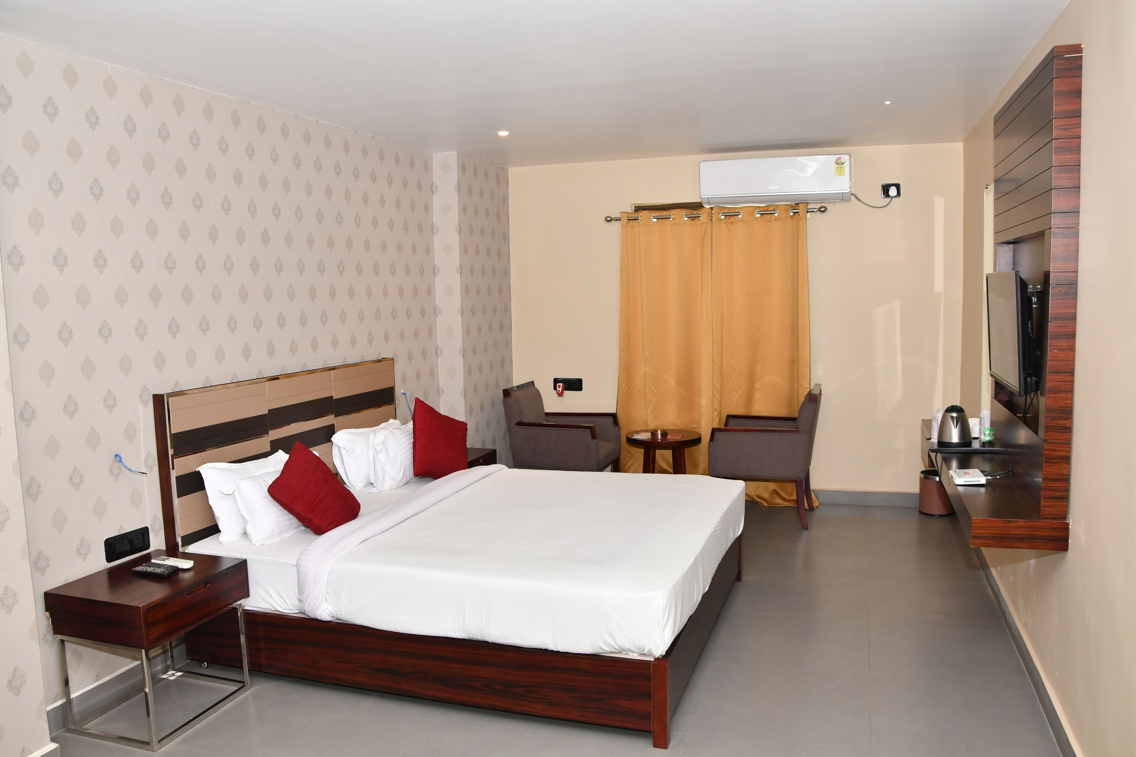 A hotel room with a large bed, a desk with a chair, a small table, a TV, and an air conditioning unit.