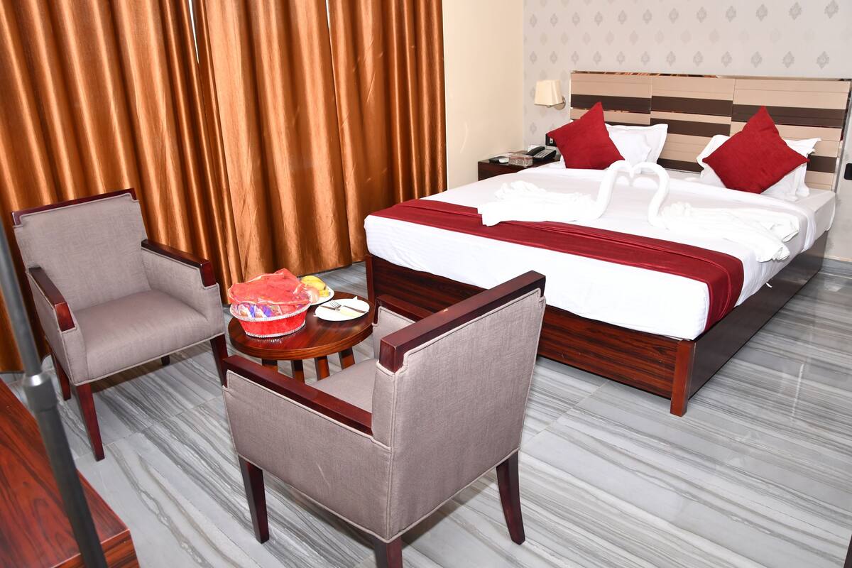 A hotel room with a large bed, two armchairs, a small round table with a cake and a plate, and a nightstand with a lamp.