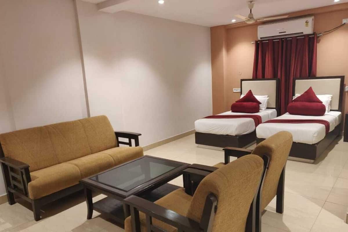 A hotel room with a sofa, a glass-top coffee table, two single beds with red pillows, and a ceiling fan.