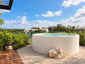 A circular pool with a wooden deck and a rocky edge, surrounded by greenery and a clear sky.