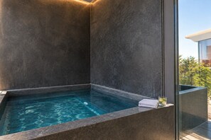 A modern bathroom with a built-in jacuzzi, dark stone walls, and a clear view of the outdoors.