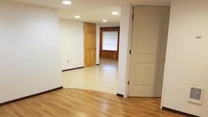 Un pasillo con suelo de madera y paredes blancas, que conduce a una habitación con una puerta y una rejilla de ventilación.