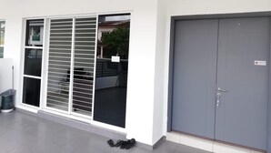 Un edificio con una puerta gris, un vidrio corredero con persianas y un contenedor de basura.