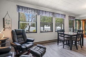 A modern living room with a leather recliner, a dining table with chairs, large windows with view curtains, and a 'live life simply' sign. - Hidden Shores Lakefront Cabin (Algoma)