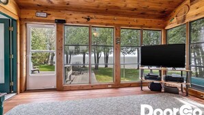 A cabin with a large TV, wooden floors, and a view of trees through large windows.