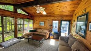 A cozy cabin interior with a wooden ceiling, a ceiling fan, a sectional sofa, a coffee table, and large windows offering a view of the forest.