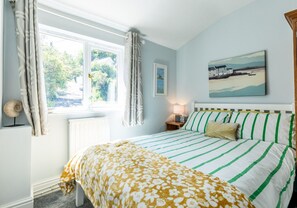 A bedroom with a bed, a window with curtains, a bedside table with a lamp, and a painting on the wall.