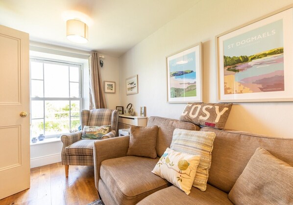 A cozy living room with a brown sofa, plaid armchair, and a window with curtains. There are framed pictures on the wall and a lamp above the sofa.