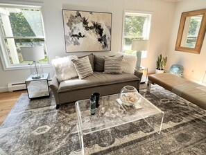 Uma sala de estar moderna com um sofá cinzento, uma mesa de centro de vidro e um grande quadro abstrato na parede.