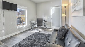 A modern living room with a black leather couch, a gray rug, a desk with a chair, a TV mounted on the wall, and a painting on the wall.
