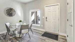 A modern dining area with a glass table and grey chairs, a white door, and a window with blinds.