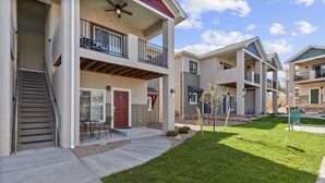 A multi-story residential building with a covered outdoor seating area, a red door, and a small lawn.