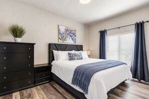 Une chambre à coucher avec un lit, une commode, une plante, un tableau et une fenêtre avec des rideaux.