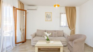 A living room with a beige sofa, a wooden coffee table, and a beige armchair. There is a window with sheer curtains, a framed picture on the wall, and an air conditioning unit. - Arvala Apartments  - Apartment with Balcony (Budva)