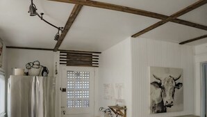 A kitchen with a stainless steel refrigerator, wooden beams on the ceiling, a white door with a window, and a wall-mounted cow painting. - Historic 1910 Estate · Sleeps 24 · Five Points South · Tea Olive & Azalea (Birmingham)