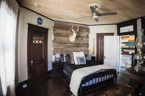 Une chambre à coucher avec une tête de lit en bois, un lit avec un cadre en métal noir, un ventilateur de plafond, une table de chevet avec une lampe et une porte avec une pancarte.